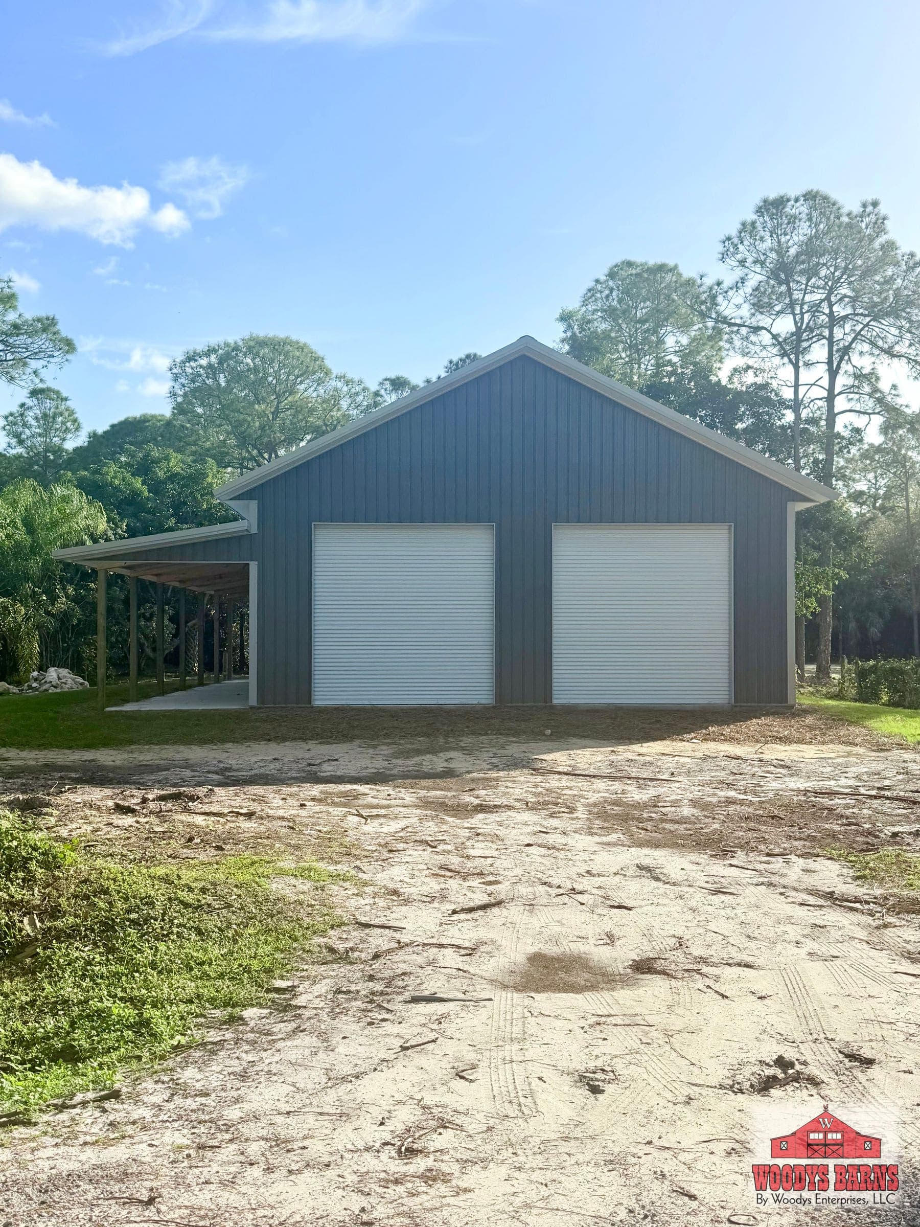 The Mack garage, a charcoal metal workshop with two white roll up doors and a covered lean to, built by Woodys Barns in Florida
